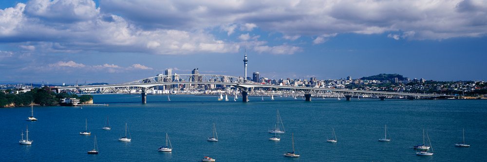City skyline of Auckland featuring the Harbour Bridge, Sky Tower, and multiple sailboats on the water under a partly cloudy blue sky, New Zealand.