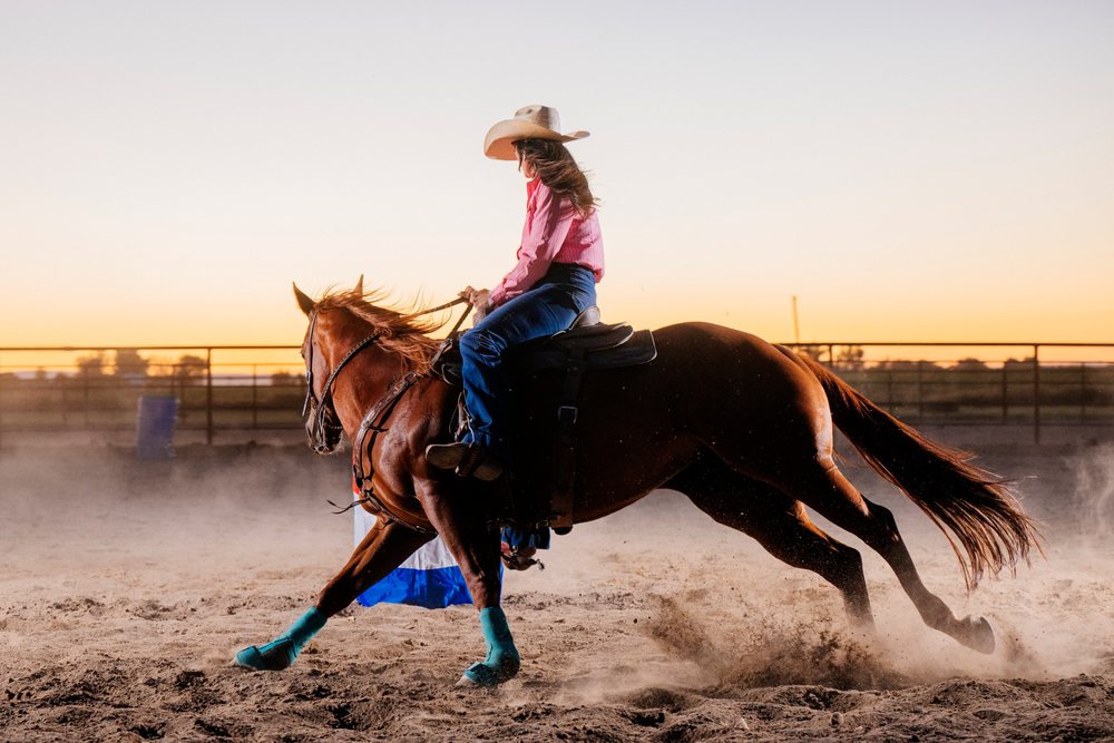 Female rider with long hair gallops a brown horse wearing turquoise leg wraps in a dusty dirt arena at sunset, Southwest USA.