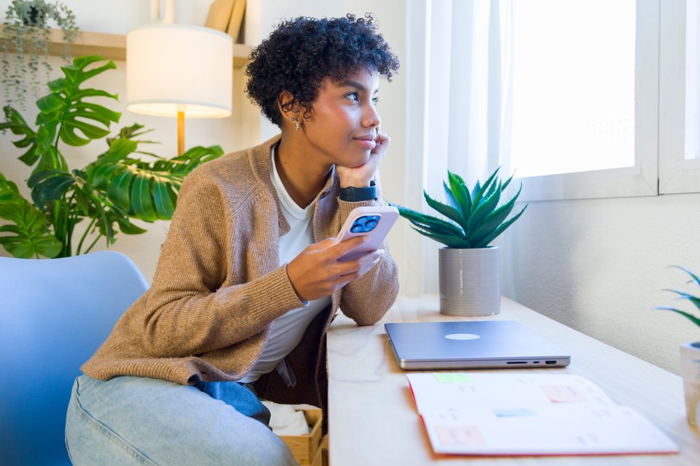 Woman holding smartphone sitting at home desk looking out window.