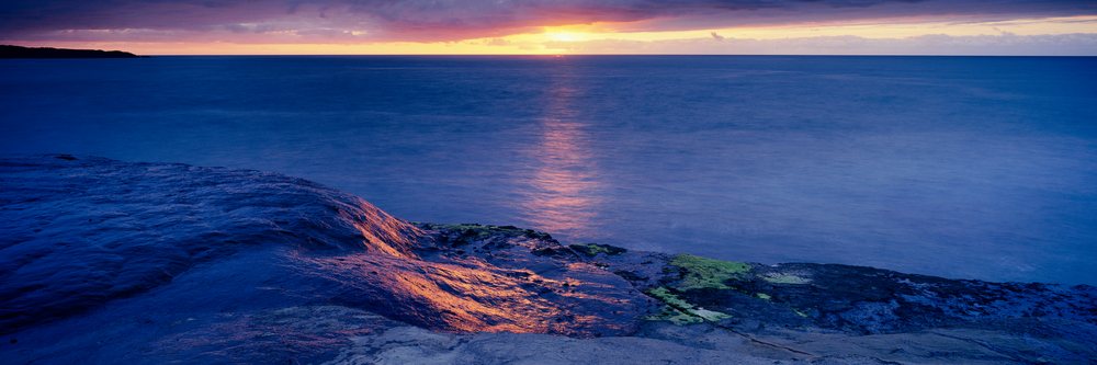 Sunset over a rocky ocean shoreline with reflection on the water and a colorful sky.