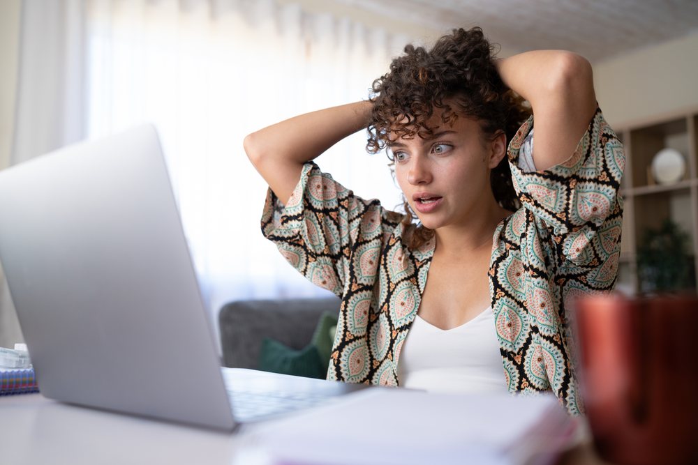 Woman with shocked expression using a laptop at a desk in a home living room in Spain during morning light.