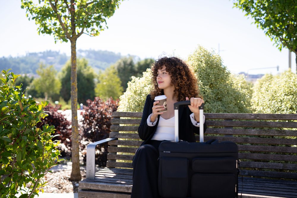 Woman with curly hair seated on a wooden bench holding a coffee cup beside a black suitcase in a sunny park setting, Spain.