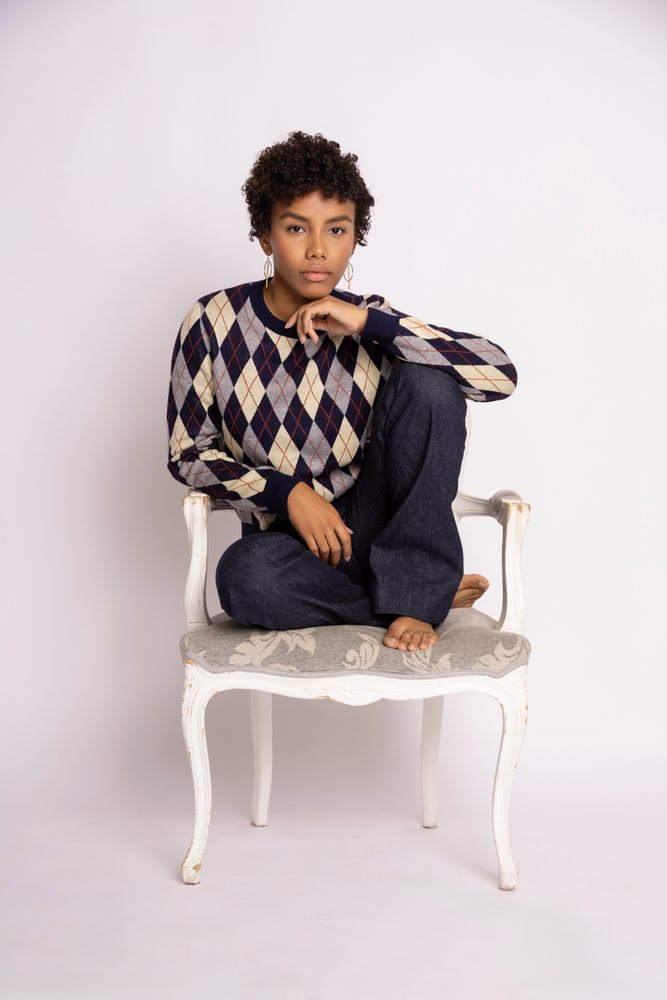 Woman sitting barefoot on a white wooden chair with decorative upholstery, wearing an argyle patterned sweater and dark trousers in a studio, fashion, young culture.