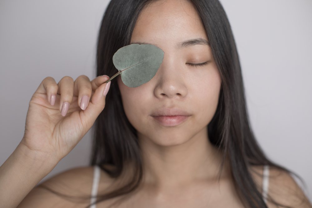 Woman with long black hair holding a green leaf over her closed eye in a calm indoor portrait, Art, emotional.