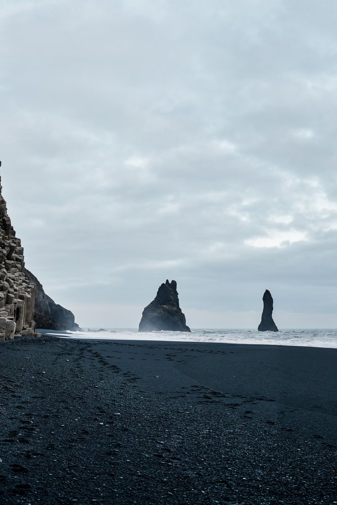 Black sand beach with footprints and basalt rock formations along the coast of Iceland under a cloudy sky.