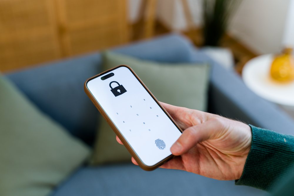 Hand holding a smartphone displaying a locked screen with numeric keypad and fingerprint icon in a living room with a blue sofa and green pillow, Spain.