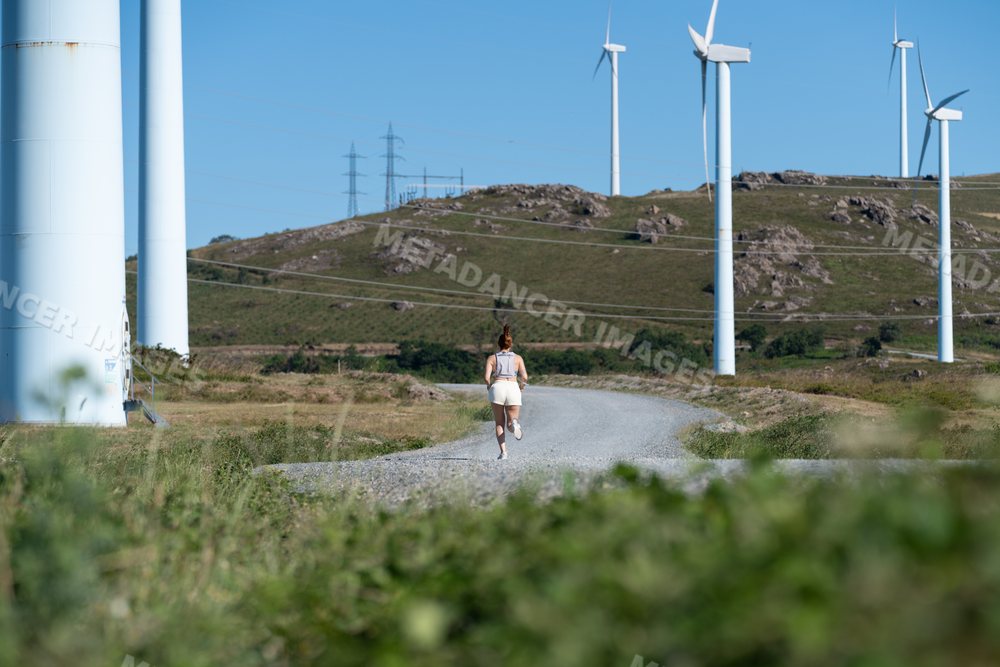 A white woman running on a gravel path near wind turbines in Galicia