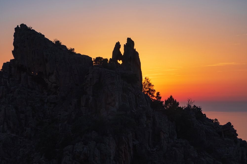 Silhouetted rock formation with heart-shaped hole at sunset, showing trees and cliffs, Travel Photography, Corsica and Sardinia, France and Italy.