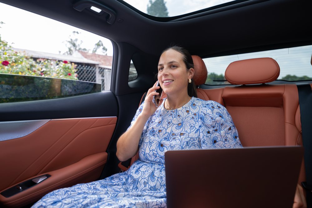 Woman in a blue floral dress sitting in the backseat of a car, smiling while talking on a mobile phone during the day, Spain.