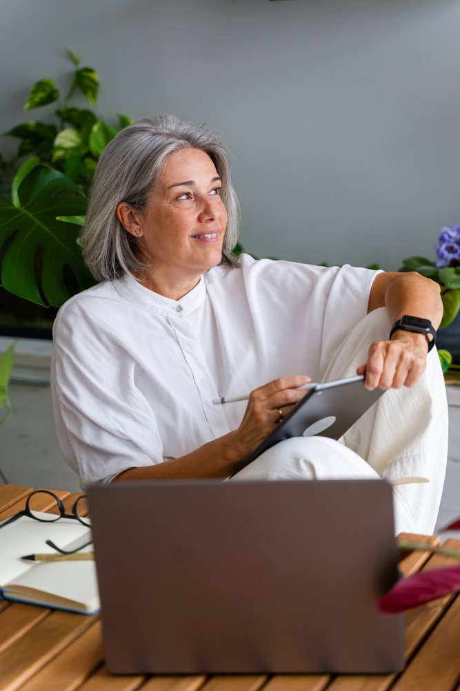 Mature woman using a digital tablet with a stylus while sitting at a wooden table with a laptop and notebook, surrounded by indoor plants in a home workspace, Spain.