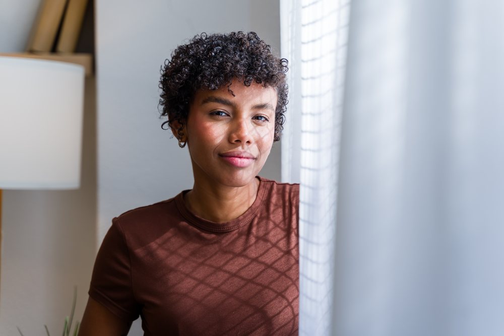 Young woman with curly black hair and earrings looks through a window indoors with natural light illuminating the modern home interior, Spain.