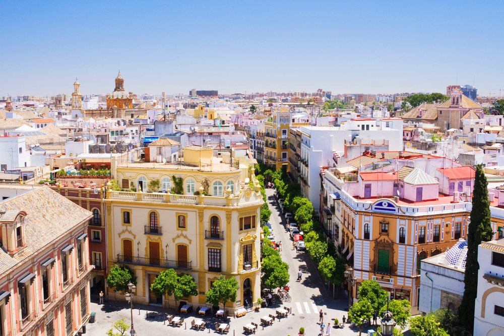 Cityscape featuring rooftops, tree-lined street, pedestrians, and outdoor tables on a clear summer day, Andalucia, Spain.