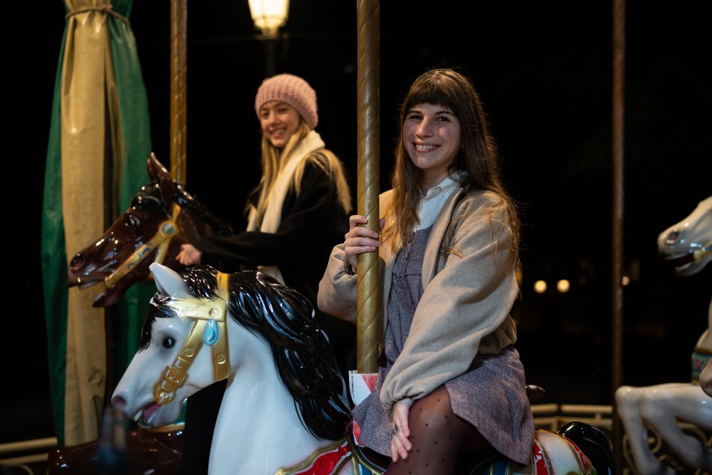 Two women smiling while riding carousel horses on a merry-go-round at night, Christmas market, Madrid, Spain.