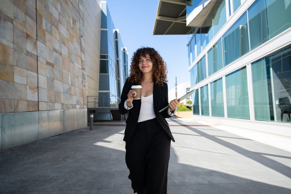 Businesswoman walking outdoors with coffee cup and folder near modern buildings, traveling executive, Spain.