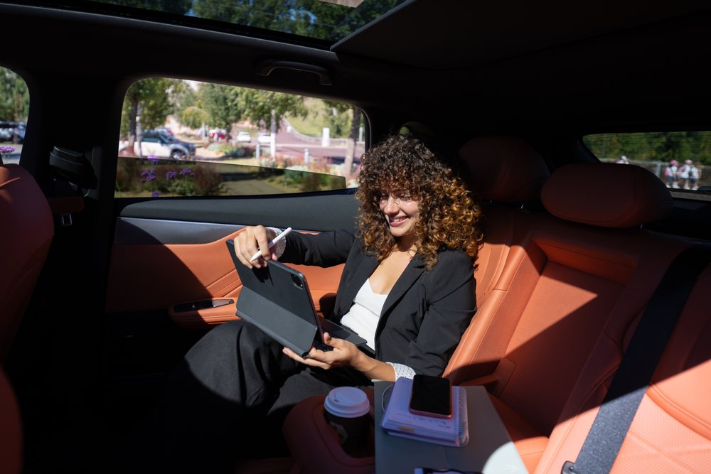 Businesswoman with curly hair using tablet and digital pen inside car with orange leather seats during remote work, Spain.