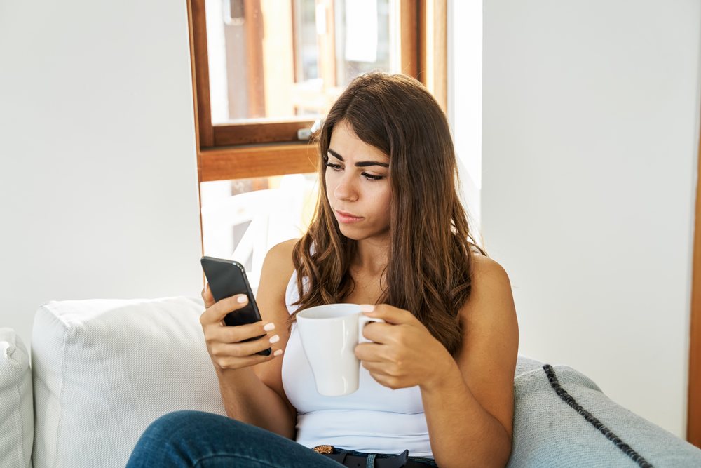 A white woman using her mobile phone while relaxing with a mug indoors