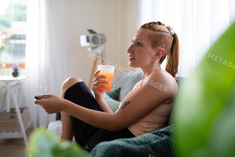 A white woman relaxing on couch with juice and remote control at home