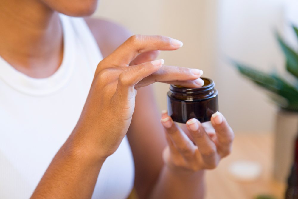 Woman holding a small open brown jar of face cream while preparing to apply skincare at home.