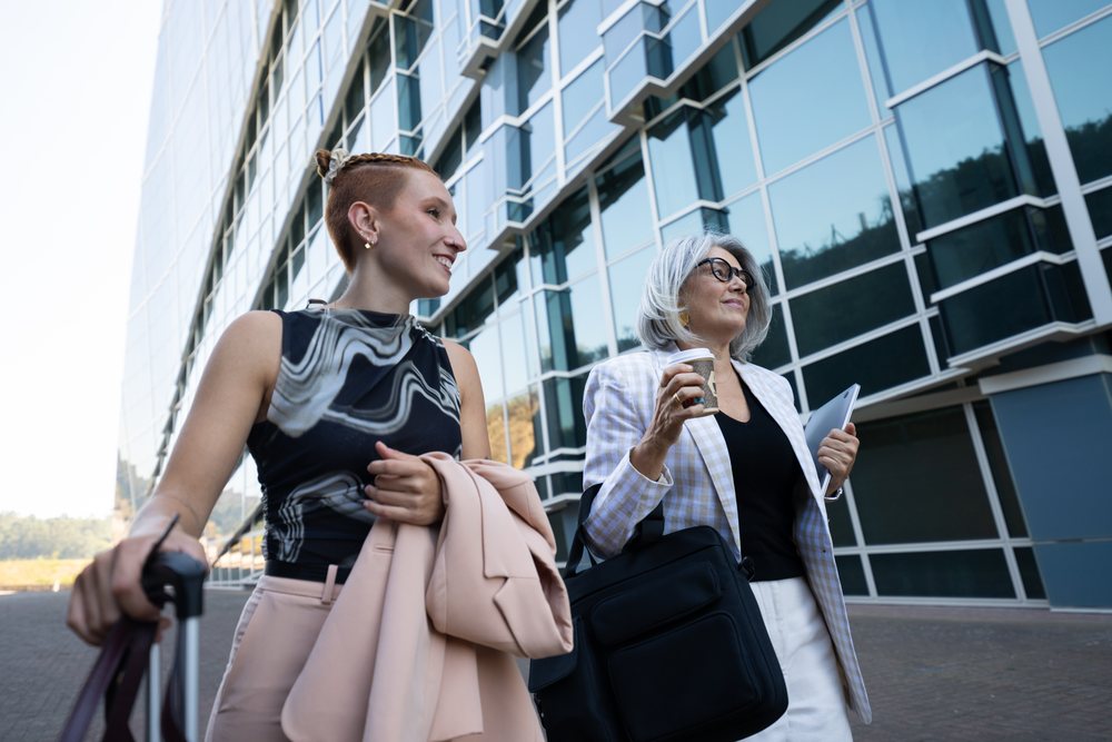 Two businesswomen walk outside a modern glass office building, one carrying a briefcase, coffee cup, and laptop, Spain.