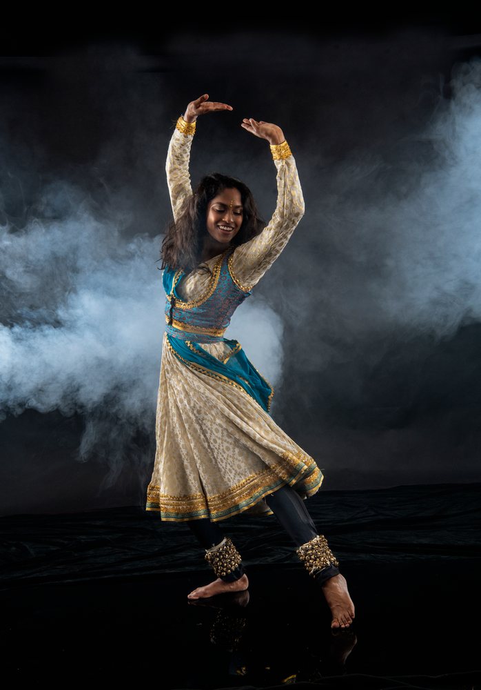 Female dancer performing Indian classical dance in costume with ankle bells and smoke on a black stage, spirituality, expression.