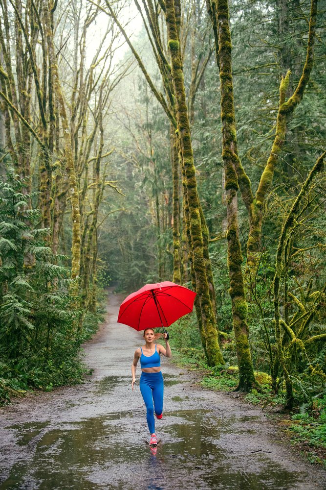 Woman running on a rainy forest trail holding a red umbrella among moss-covered trees and puddles, USA.