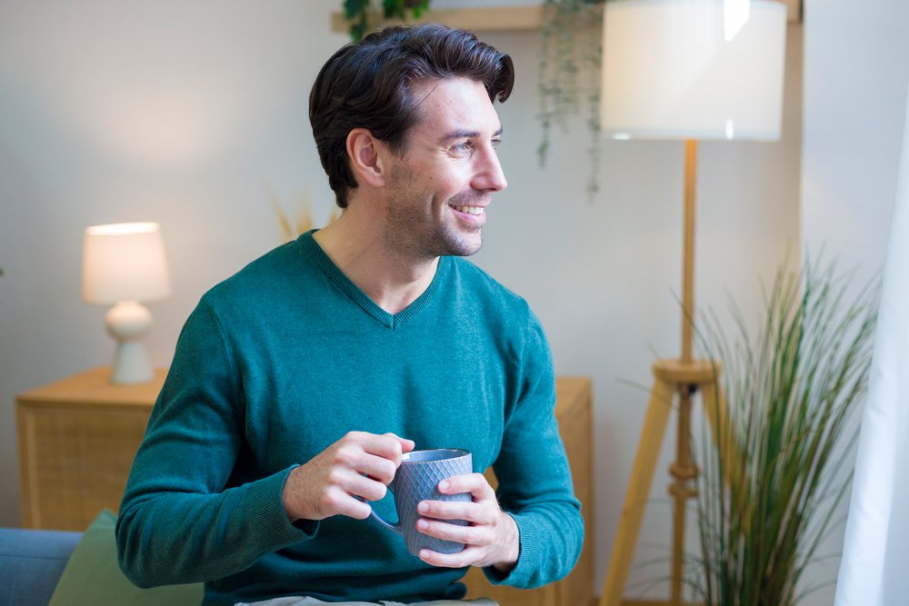 Man in a casual sweater smiles while holding a coffee cup indoors in a modern living room setting, technology, young adult, home.