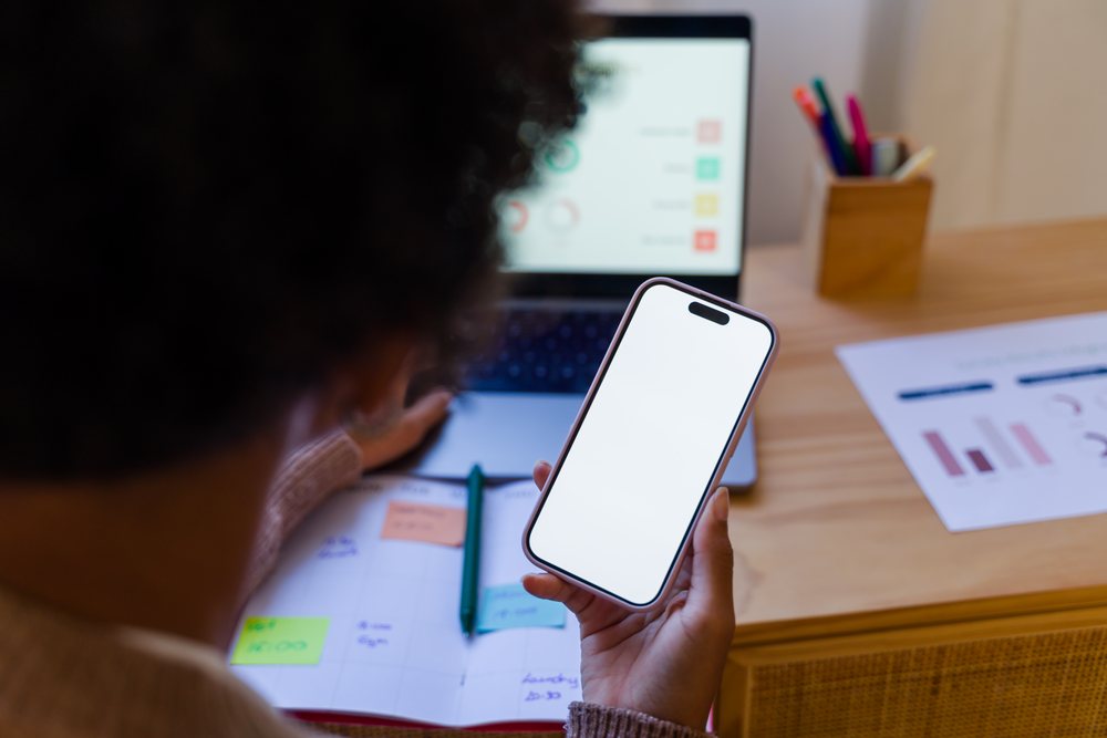 Person with curly hair holding a mobile phone with blank screen at a wooden desk with laptop, notebook, pen, and colorful pens in an office workspace, Spain.