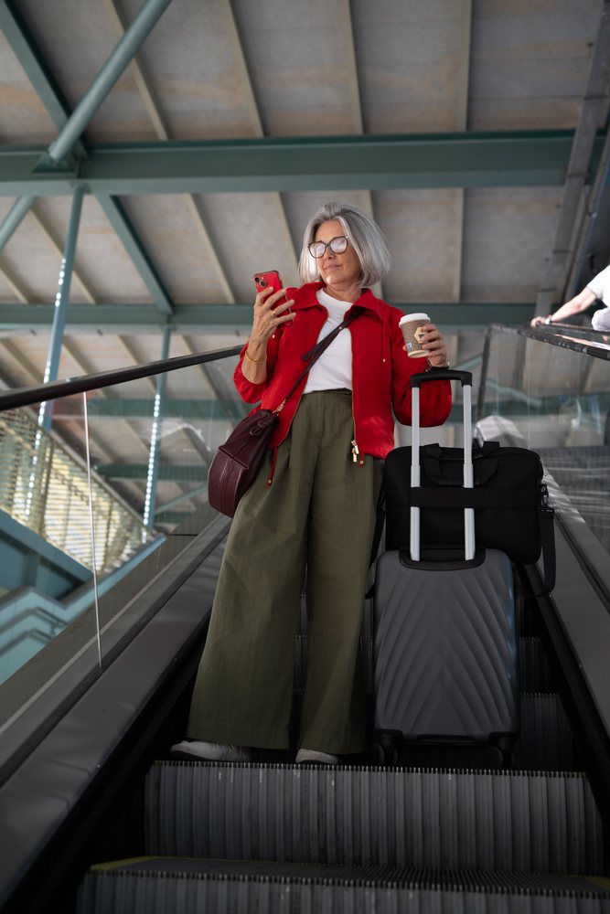 Woman with gray hair and glasses stands on an airport escalator holding coffee and looking at her smartphone, Spain.