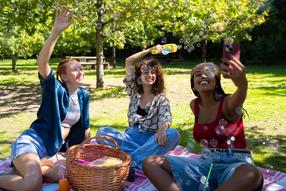 Three young women enjoying a sunny picnic in a park, blowing bubbles and taking a selfie on a blanket with a picnic basket nearby, Spain.