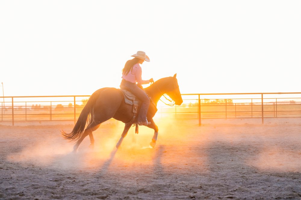 Woman wearing a cowboy hat rides a horse kicking up dust in a fenced outdoor arena at sunset, Southwest USA.