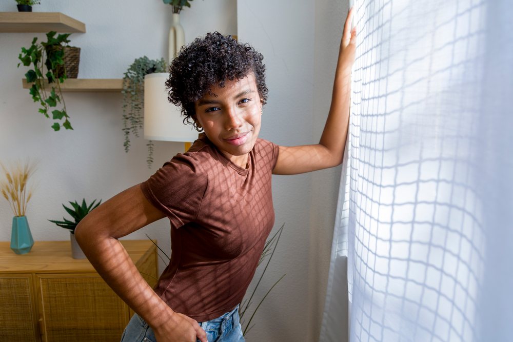 Young woman with curly hair wearing a brown shirt and blue jeans stands by a window with white curtains in a home interior with natural light.