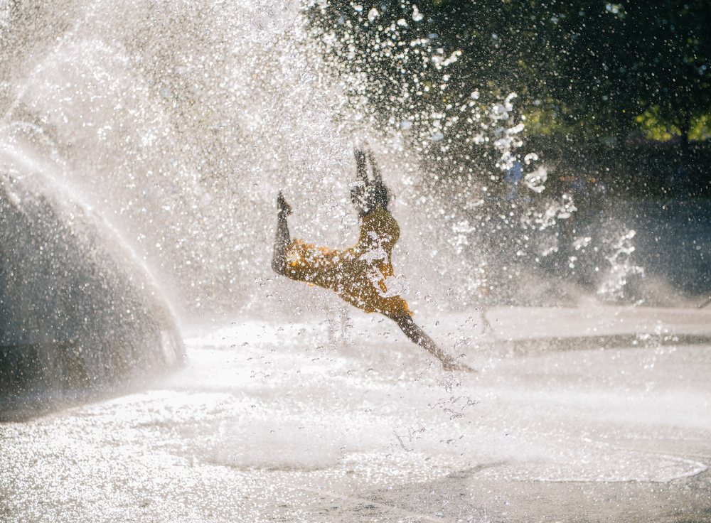 Child wearing yellow shirt playing and splashing water at an outdoor fountain in summer with sunlight and green foliage, USA.