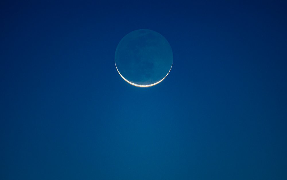 Crescent moon with faint full lunar surface visible in clear blue night sky over, Seattle, USA.