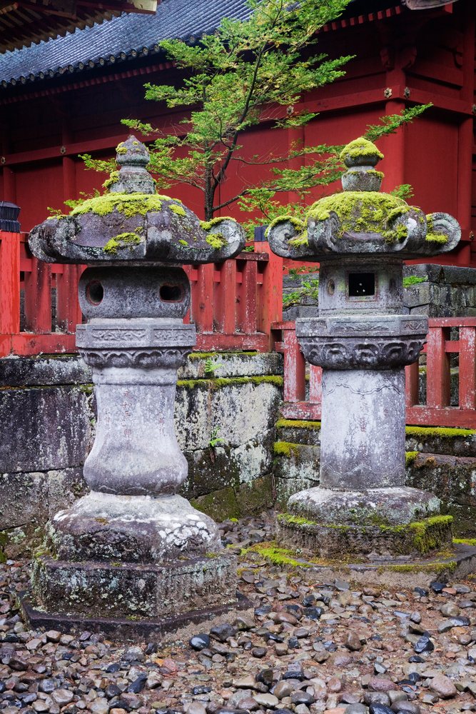 Stone lanterns covered in moss stand on rocky ground near a red wooden fence at Toshogu Shrine in Nikko National Park, Honshu, Japan.