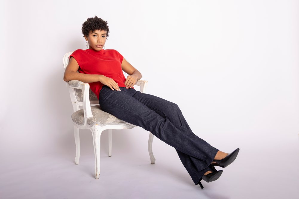 Woman with curly hair wearing a red top and black fitted pants sitting on a white vintage chair in a studio with white background, fashion, clothes, young culture.