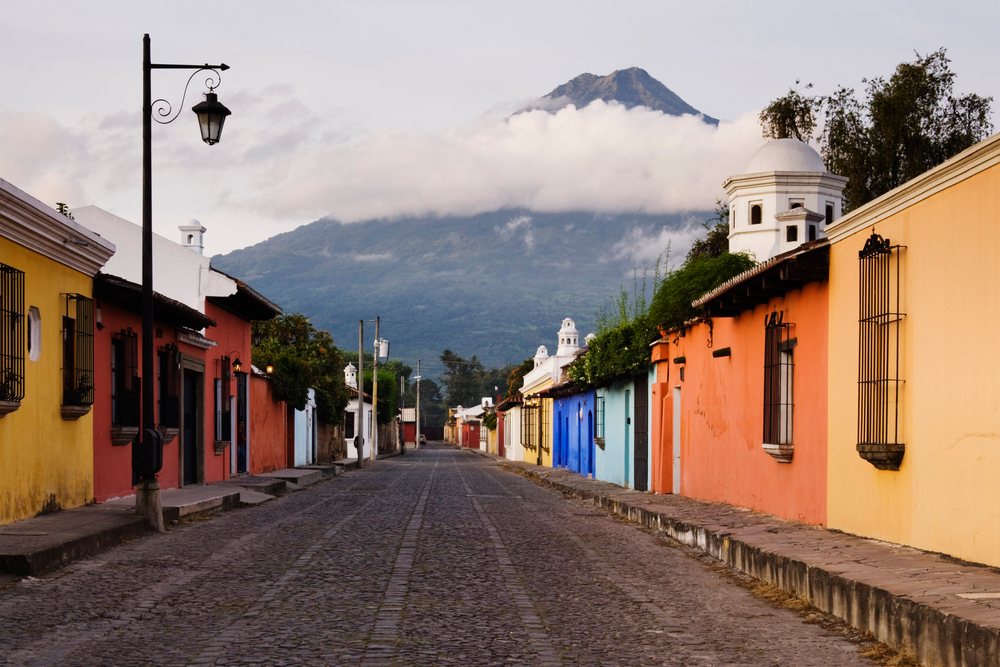 Volcano rises above an empty cobblestone street lined with colorful colonial houses Guatemala under a cloudy sky, Antigua.