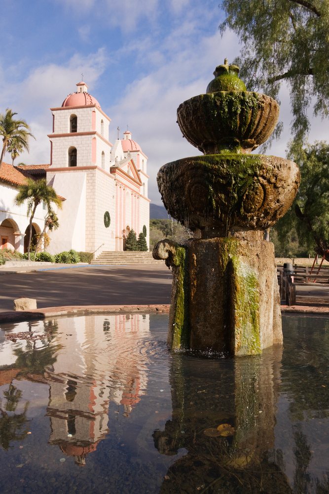Historic Spanish colonial Santa Barbara Mission with moss-covered fountain and water reflection on a sunny day, California, USA.