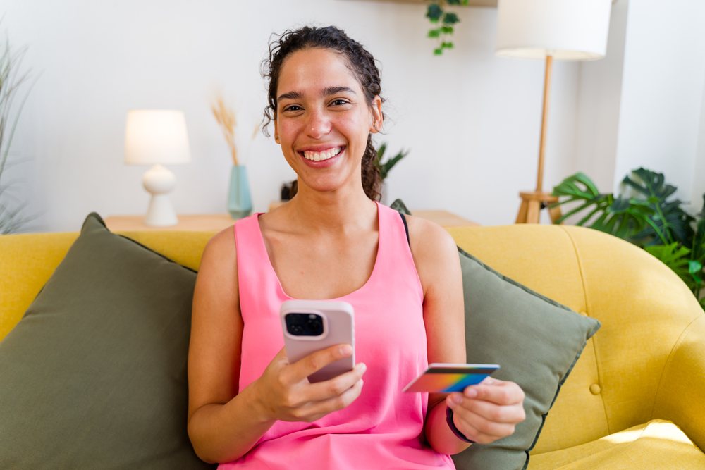 A smiling woman wearing a pink top holds a mobile phone and credit card while sitting on a yellow couch in a home interior, Spain.