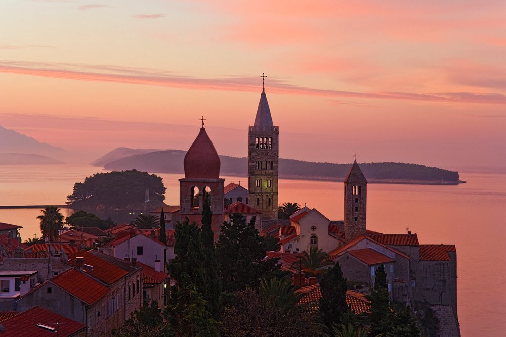 Town of Rab with church towers and red tile roofs overlooking sea and islands under a colorful sky, Dawn, Rab Island, Croatia.