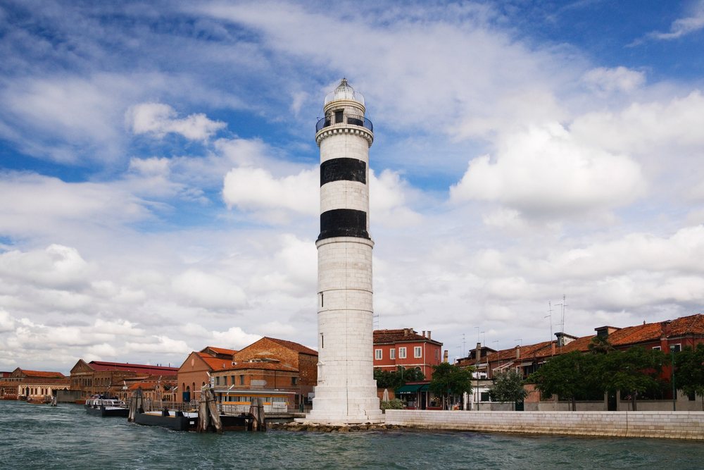 Lighthouse with black stripes standing beside a canal with brick buildings and red tiled roofs in Murano, Italy under a partly cloudy sky, Venice.