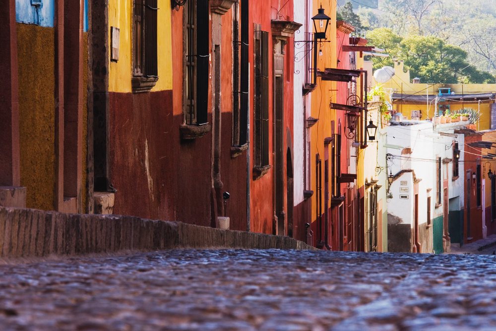 Colorful buildings line a cobblestone street with street lamps and mountain views, Guanajuato, Mexico.