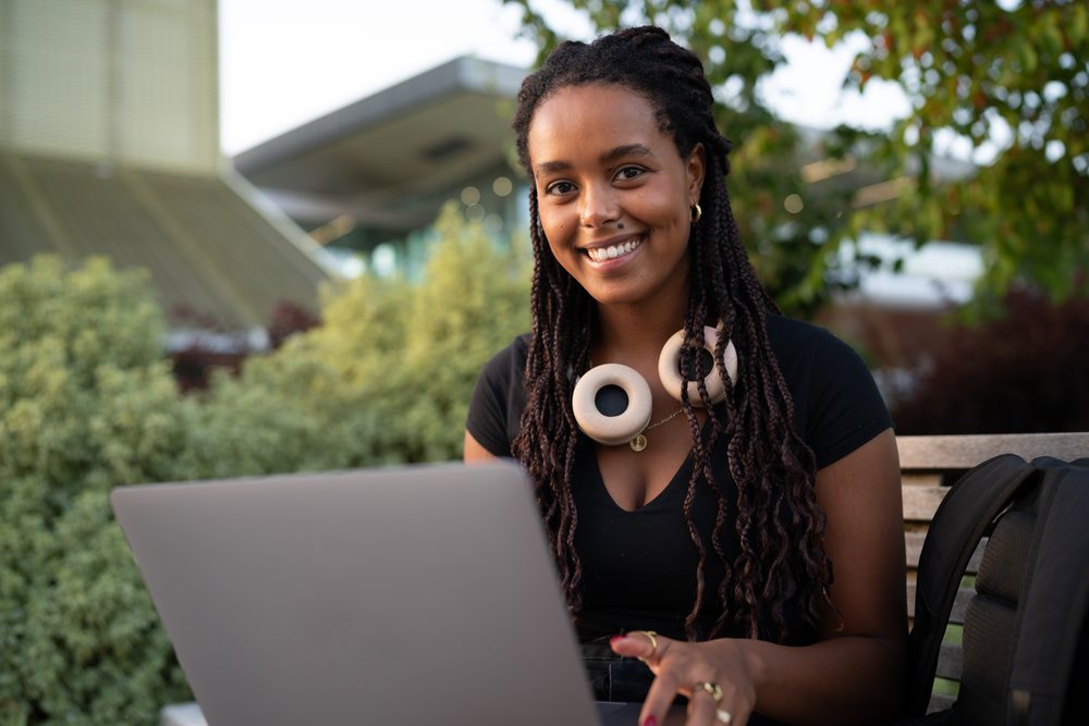 A smiling woman with braided hair works on a laptop while sitting on a park bench surrounded by greenery, urban, Santiago De Compostela, Spain.