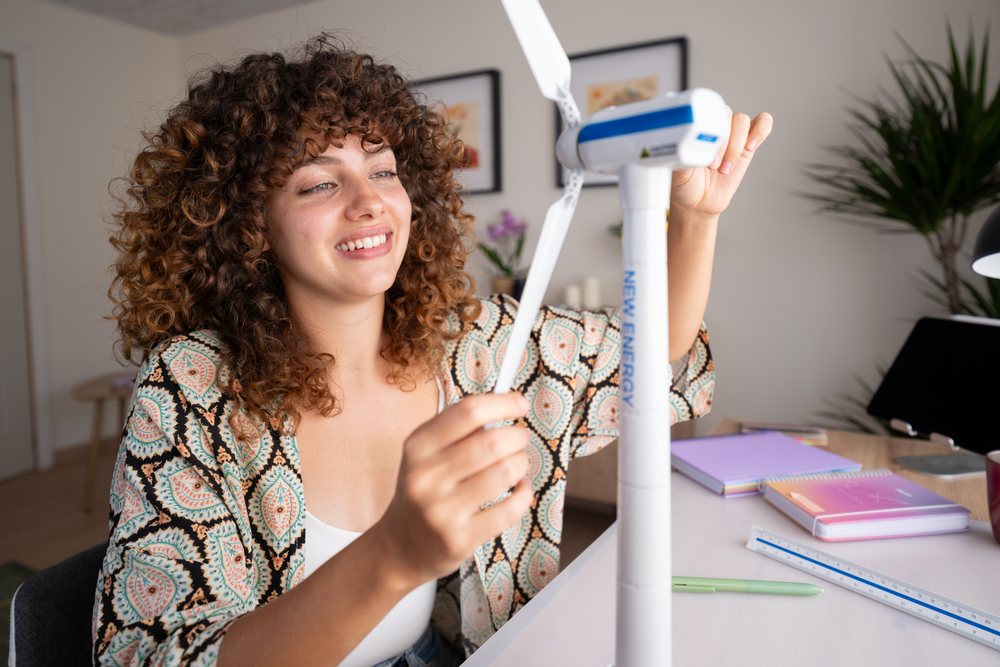 Woman with curly hair assembling a white and blue wind turbine model at a desk with notebooks, ruler, and pen in a home setting, Spain.