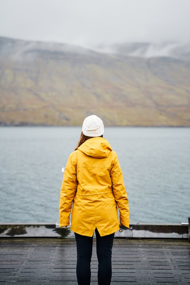 A woman wearing a yellow jacket and white knit hat stands on a wooden dock looking at a lake surrounded by misty mountains, Iceland.