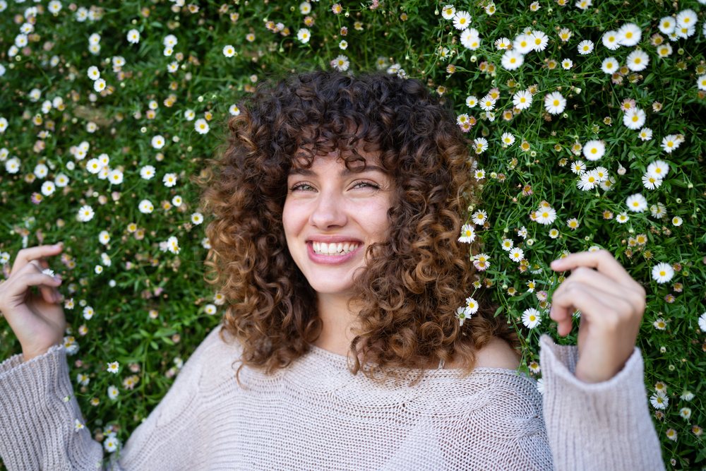 Smiling woman with curly hair lays in a field of daisies and greenery outdoors, home story, influencer, Spain.