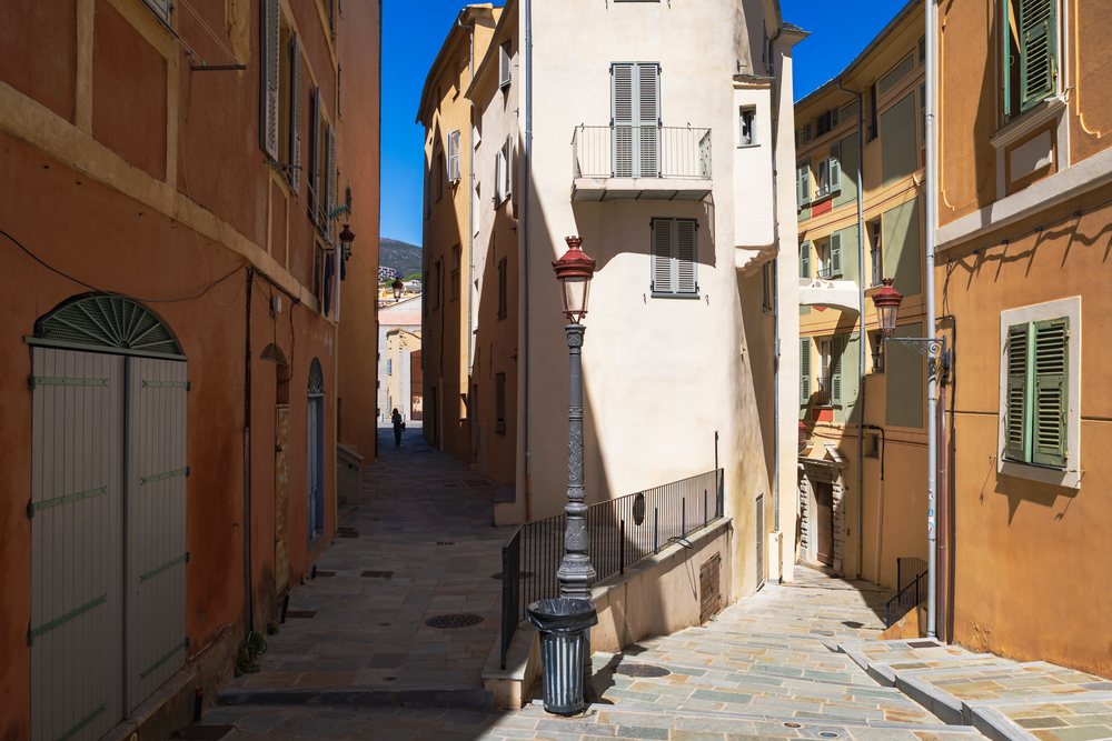 Narrow sunlit stone-paved street with yellow and cream buildings and green shutters, Travel Photography, Bastia, Corsica, France.