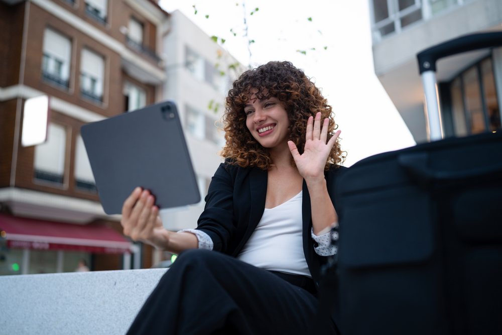 Young woman seated outdoors in Madrid smiling and waving during a video call on a tablet.