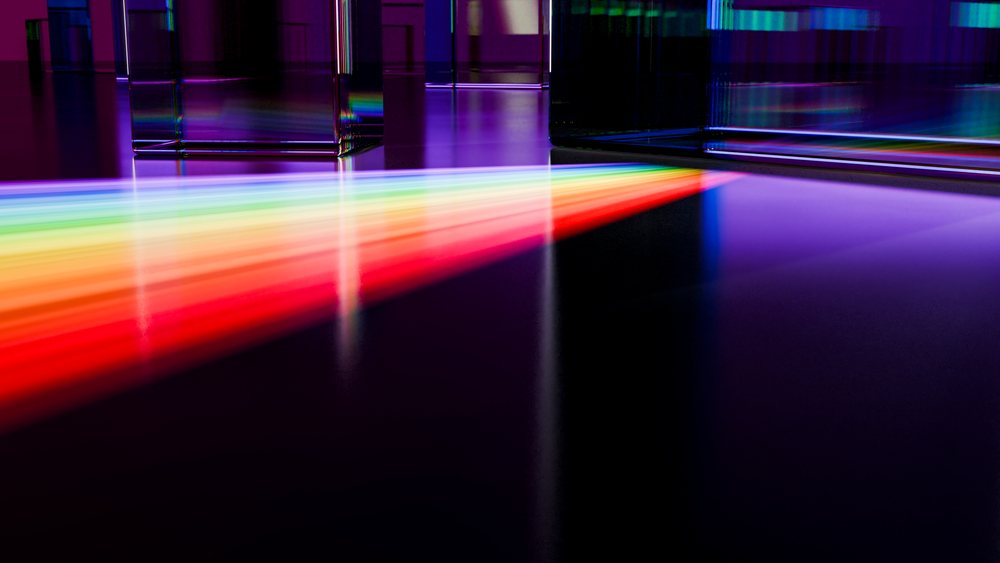Glass prisms refract and disperse a light beam into a colorful spectrum reflected on a dark surface.