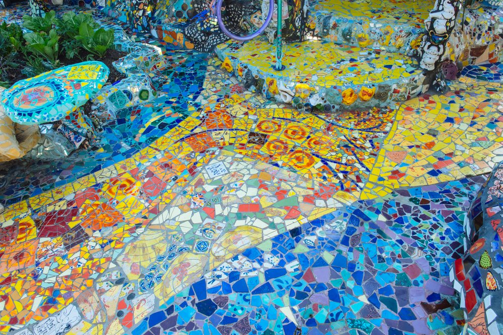 Colorful broken ceramic tiles form an outdoor mosaic art with green plants visible beside it in a garden setting, CA, USA.