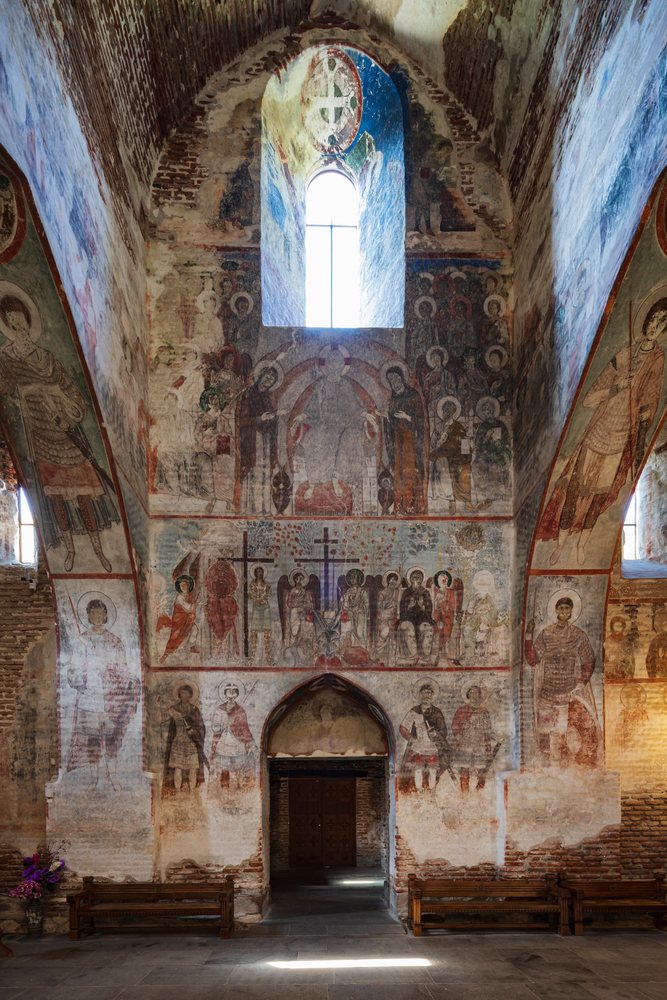 Interior of Tsagveri Timothesubvani Monastery showing 11th century frescoes on brick walls, wooden benches, arched window, and stone floor, Georgia.